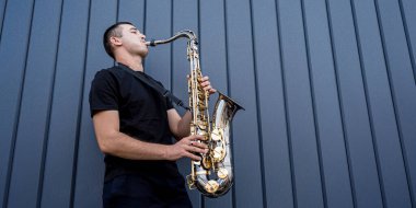 Genç sokak müzisyeni büyük mavi duvarın yanında saksafon çalıyor.