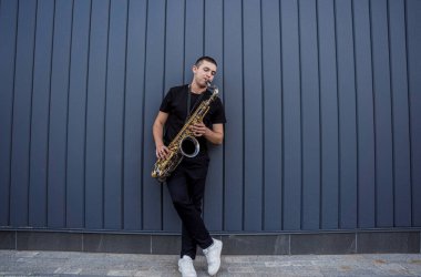 Genç sokak müzisyeni büyük mavi duvarın yanında saksafon çalıyor.