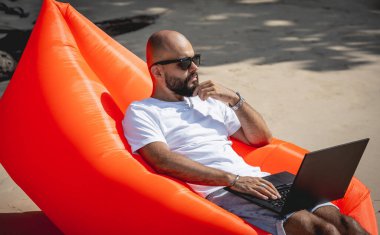 Genç adam şişme bir koltukta otururken kumsalda dizüstü bilgisayarıyla çalışıyor.