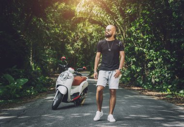 Şık bir genç adam ve motosikleti ormanda yolda.