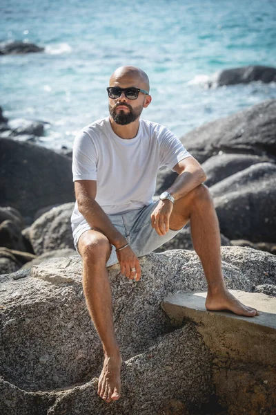 Genç adam kumsalda dinlenirken bir taşın üzerinde oturuyor.