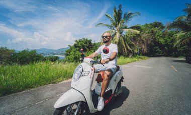 Şık bir genç adam deniz kenarında motosiklet sürüyor ve palmiye ağaçları.