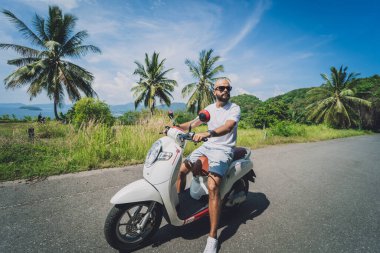 Şık bir genç adam deniz kenarında motosiklet sürüyor ve palmiye ağaçları.