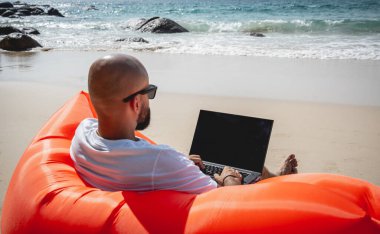 Genç adam şişme bir koltukta otururken kumsalda dizüstü bilgisayarıyla çalışıyor.