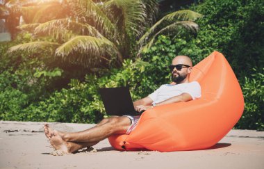 Genç adam şişme bir koltukta otururken kumsalda dizüstü bilgisayarıyla çalışıyor.