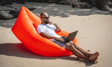 Genç adam şişme bir koltukta otururken kumsalda dizüstü bilgisayarıyla çalışıyor.