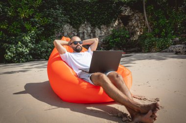 Genç adam şişme bir koltukta otururken kumsalda dizüstü bilgisayarıyla çalışıyor.