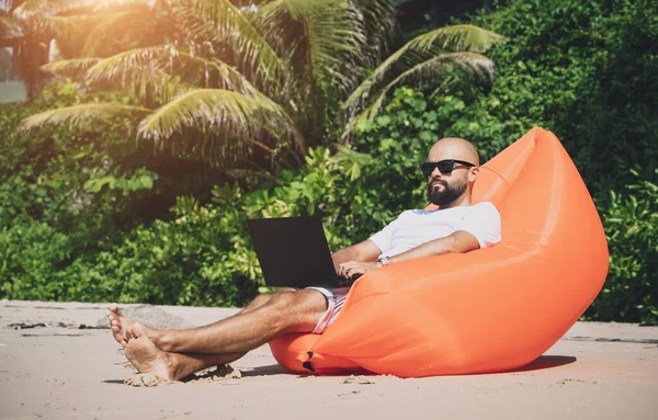 Genç adam şişme bir koltukta otururken kumsalda dizüstü bilgisayarıyla çalışıyor.