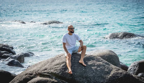 Genç adam kumsalda dinlenirken bir taşın üzerinde oturuyor.