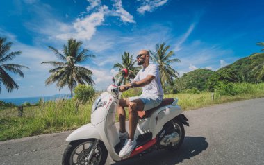 Şık bir genç adam deniz kenarında motosiklet sürüyor ve palmiye ağaçları.