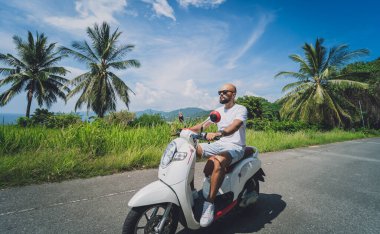 Şık bir genç adam deniz kenarında motosiklet sürüyor ve palmiye ağaçları.