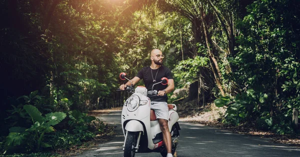 Şık bir genç adam ve motosikleti ormanda yolda.