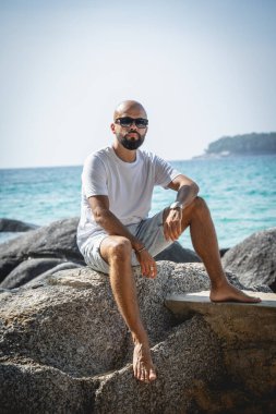 Genç adam kumsalda dinlenirken bir taşın üzerinde oturuyor.