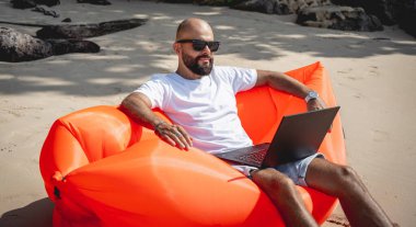 Genç adam şişme bir koltukta otururken kumsalda dizüstü bilgisayarıyla çalışıyor.