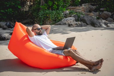 Genç adam şişme bir koltukta otururken kumsalda dizüstü bilgisayarıyla çalışıyor.