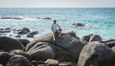 Genç adam kumsalda dinlenirken bir taşın üzerinde oturuyor.