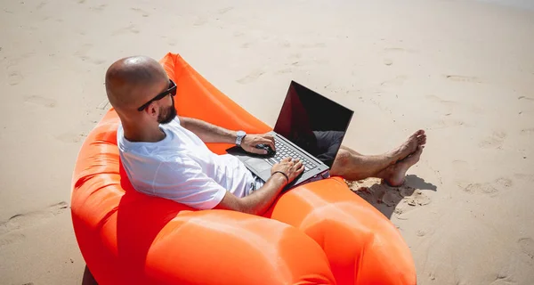 Νεαρός που δουλεύει στο λάπτοπ του στην παραλία ενώ κάθεται σε έναν φουσκωτό καναπέ. — Φωτογραφία Αρχείου