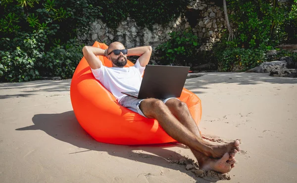 Genç adam şişme bir koltukta otururken kumsalda dizüstü bilgisayarıyla çalışıyor.