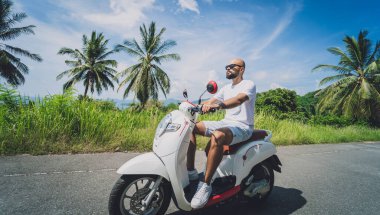 Şık bir genç adam deniz kenarında motosiklet sürüyor ve palmiye ağaçları.