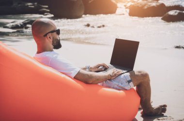 Young man working on his laptop on the beach while sitting on an inflatable sofa