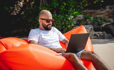 Genç adam şişme bir koltukta otururken kumsalda dizüstü bilgisayarıyla çalışıyor.