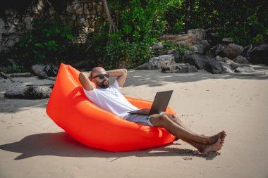 Genç adam şişme bir koltukta otururken kumsalda dizüstü bilgisayarıyla çalışıyor.
