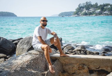 Genç adam kumsalda dinlenirken bir taşın üzerinde oturuyor.