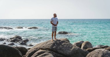 Genç adam sahilde dinlenirken bir taşın üzerinde duruyor.