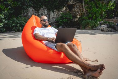 Genç adam şişme bir koltukta otururken kumsalda dizüstü bilgisayarıyla çalışıyor.