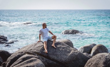 Genç adam kumsalda dinlenirken bir taşın üzerinde oturuyor.