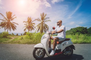 Şık bir genç adam deniz kenarında motosiklet sürüyor ve palmiye ağaçları.
