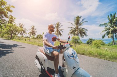 Şık bir genç adam deniz kenarında motosiklet sürüyor ve palmiye ağaçları.