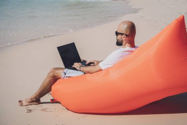 Genç adam şişme bir koltukta otururken kumsalda dizüstü bilgisayarıyla çalışıyor..