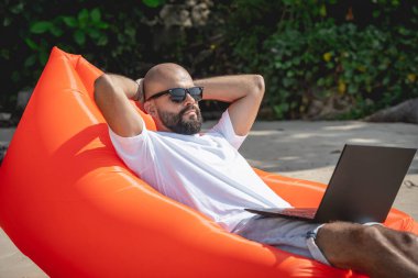 Genç adam şişme bir koltukta otururken kumsalda dizüstü bilgisayarıyla çalışıyor..