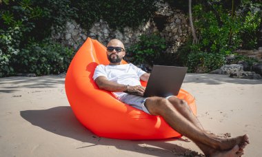 Genç adam şişme bir koltukta otururken kumsalda dizüstü bilgisayarıyla çalışıyor..