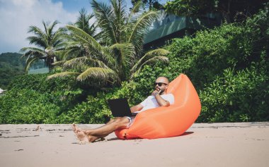 Genç adam şişme bir koltukta otururken kumsalda dizüstü bilgisayarıyla çalışıyor..