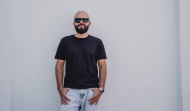 Beyaz bir duvarın arkasında siyah tişört giyen sakallı bir erkek model..