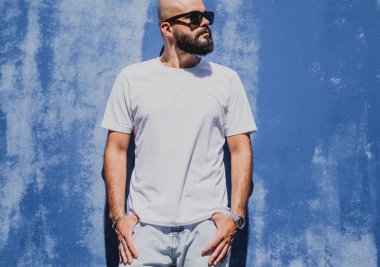 Mavi bir duvarın arkasında beyaz tişört giyen sakallı bir erkek model..