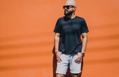 Turuncu bir duvarın arkasında siyah tişört giyen sakallı bir erkek model..