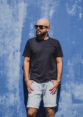 Mavi bir duvarın arkasında siyah, boş tişört giyen sakallı bir erkek model..