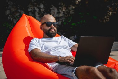 Genç adam şişme bir koltukta otururken kumsalda dizüstü bilgisayarıyla çalışıyor..