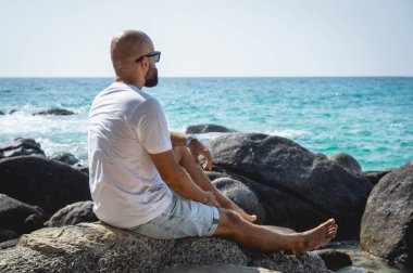 Genç adam kumsalda dinlenirken bir taşın üzerinde oturuyor..