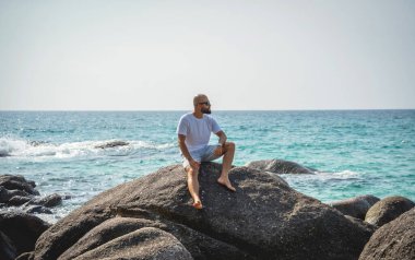 Genç adam kumsalda dinlenirken bir taşın üzerinde oturuyor..