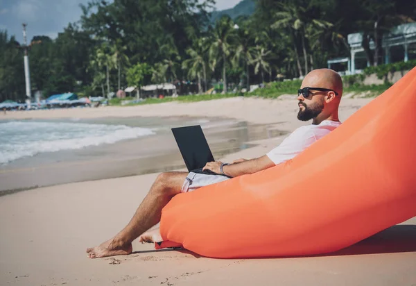 Genç adam şişme bir koltukta otururken kumsalda dizüstü bilgisayarıyla çalışıyor..