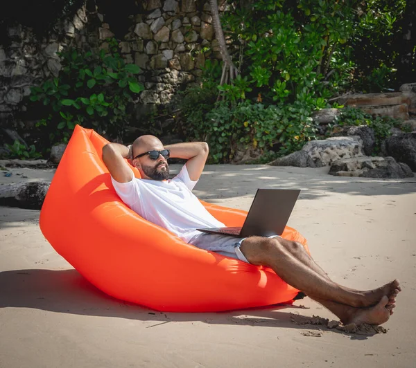 Genç adam şişme bir koltukta otururken kumsalda dizüstü bilgisayarıyla çalışıyor..