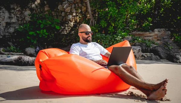 Genç adam şişme bir koltukta otururken kumsalda dizüstü bilgisayarıyla çalışıyor..