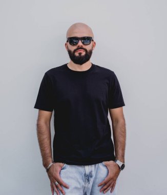 Beyaz bir duvarın arkasında siyah tişört giyen sakallı bir erkek model..