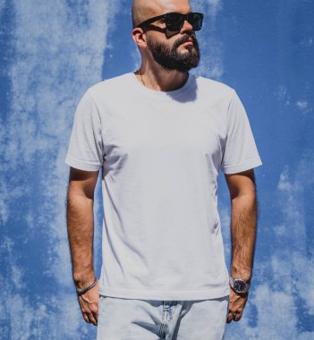 Mavi bir duvarın arkasında beyaz tişört giyen sakallı bir erkek model..