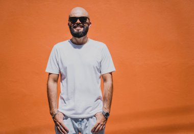 Turuncu bir duvarın arkasında beyaz tişört giyen sakallı bir erkek model..