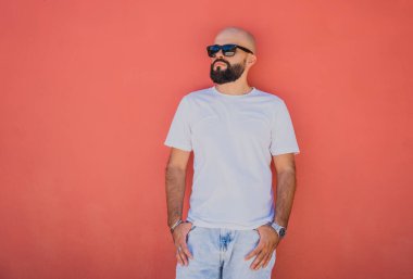 Turuncu bir duvarın arkasında beyaz tişört giyen sakallı bir erkek model..